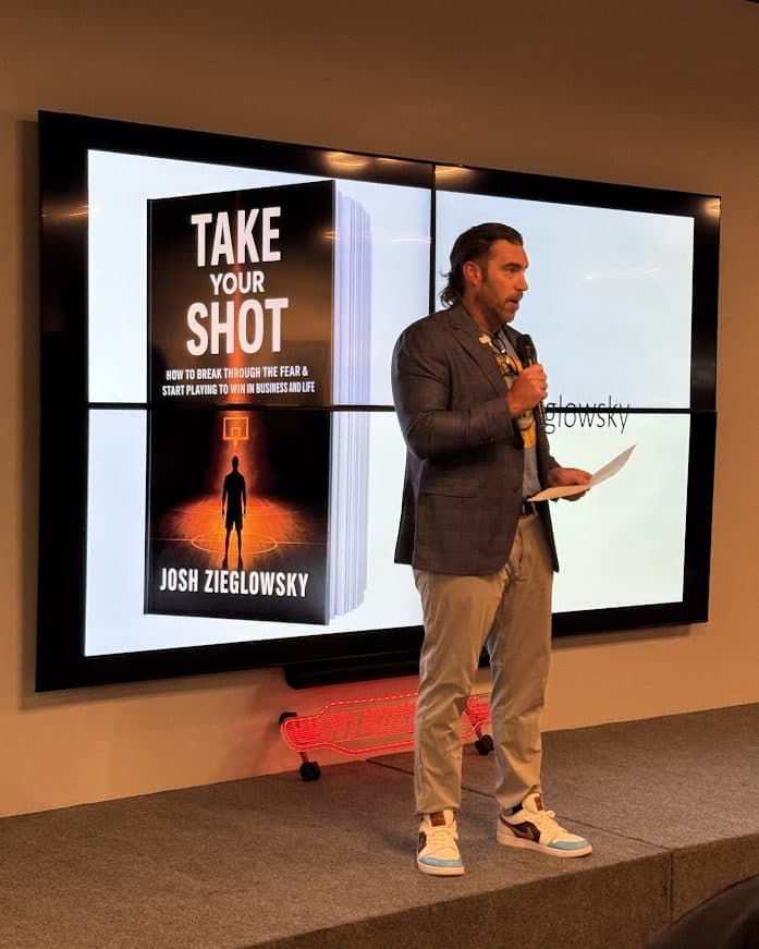 Josh Zieglowsky speaking on stage, with the Take Your Shot cover on screen behind him